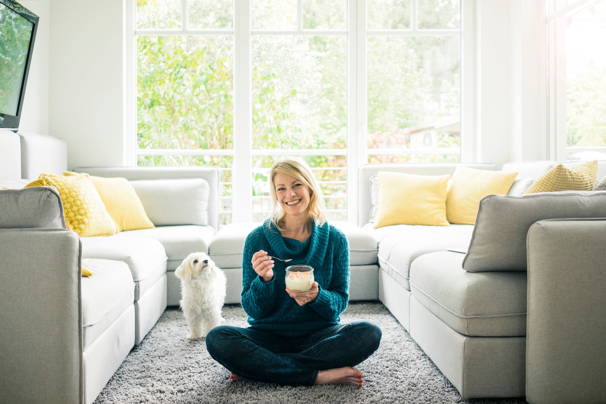 Mulher comendo iogurte em sala de estar enquanto cãozinho branco olha para ela